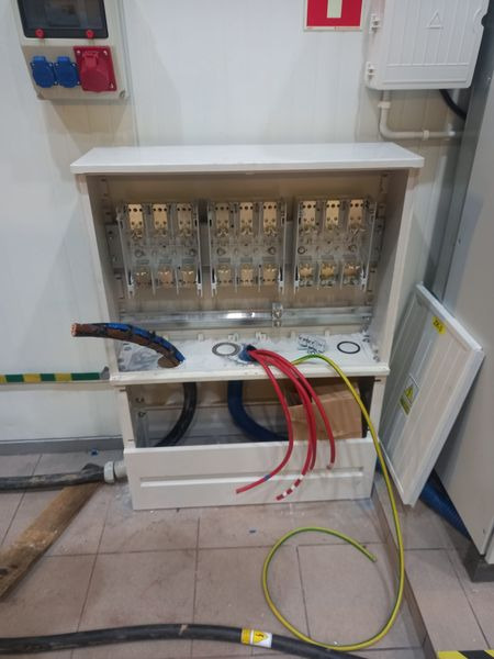 Otwarta skrzynka elektryczna z widocznymi przewodami w kolorach czerwonym, żółto-zielonym i niebieskim, oraz grubym czarnym kablem w podłodze, w trakcie instalacji w pomieszczeniu przemysłowym.