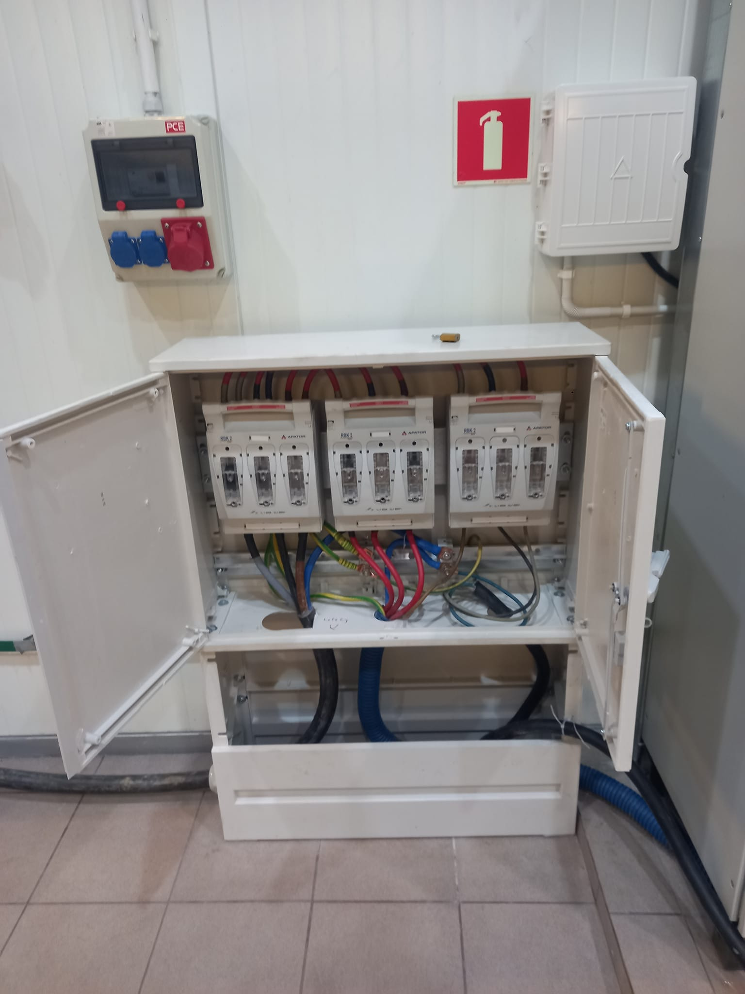 Otwarta skrzynka elektryczna z trzema bezpiecznikami RBK2, widoczne przewody kolorowe, dodatkowa skrzynka z gniazdami siłowymi na ścianie, obok znak gaśnicy.