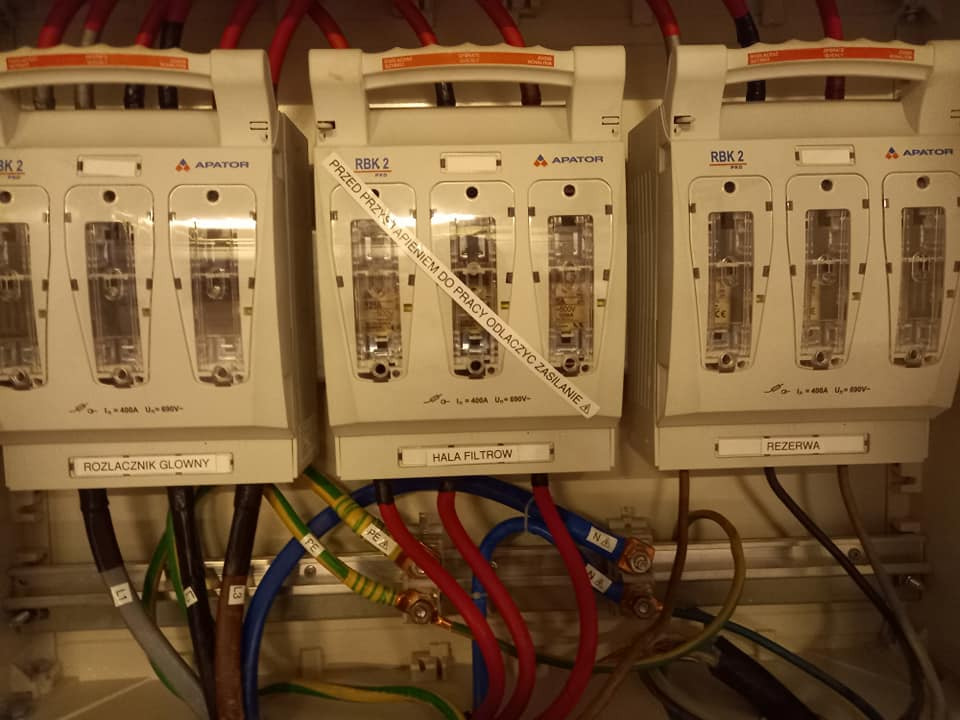 Rozdzielnica elektryczna z trzema modułami RBK2 firmy Apator, okablowanie zasilające, etykieta ostrzegawcza o odłączeniu zasilania przed pracą, moduły oznaczone jako 'Rozłącznik Główny', 'Hala...