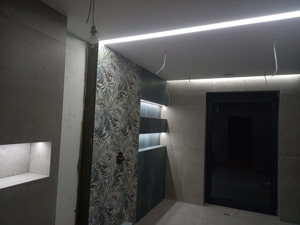 Wnętrze łazienki w trakcie wykańczania, z podświetlanymi wnękami, dekoracyjną tapetą w liście i liniowym oświetleniem LED pod sufitem, widoczne wystające przewody elektryczne.