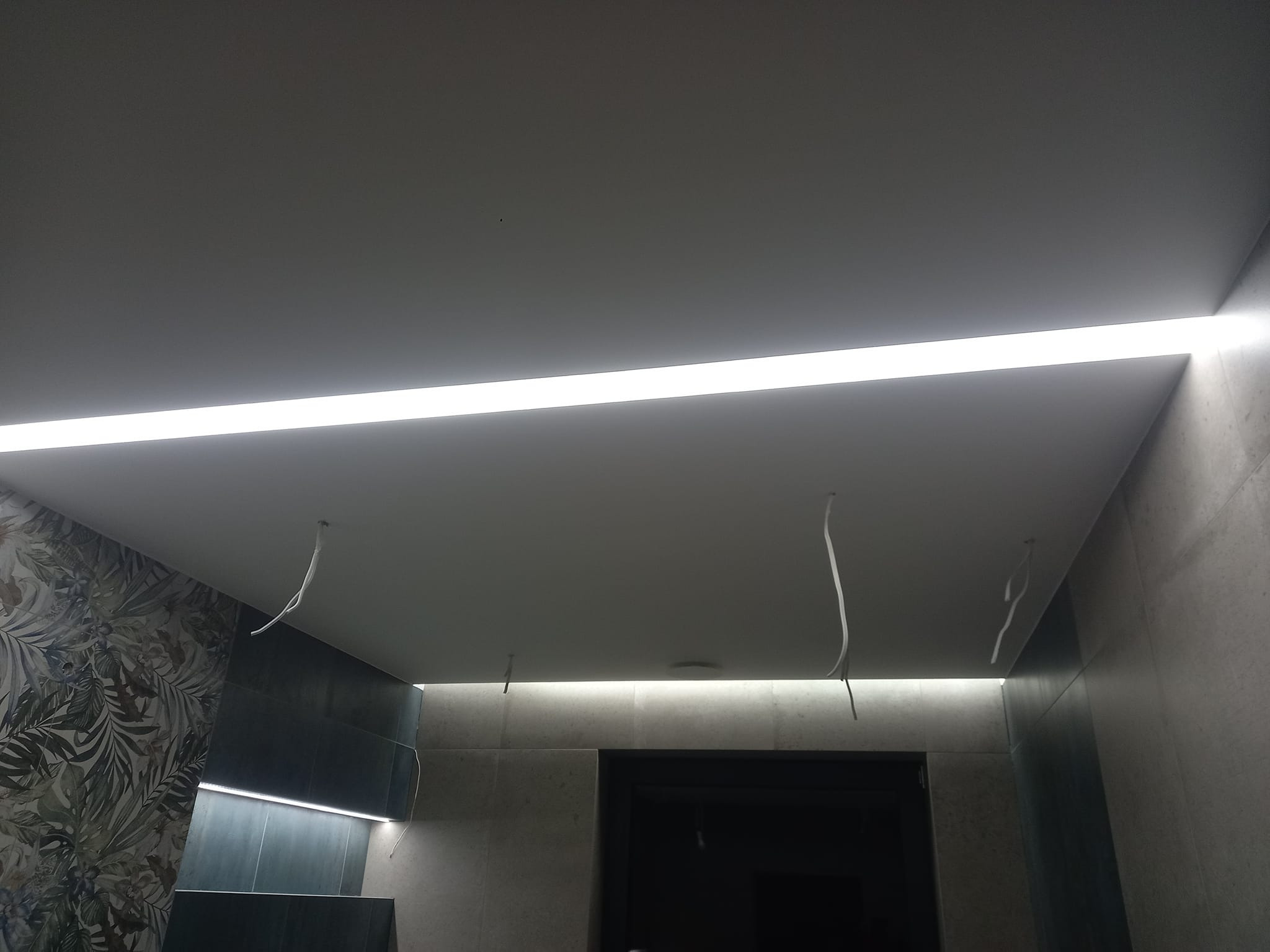 Podwieszany sufit z oświetleniem liniowym LED w łazience z dekoracyjną ścianą z motywem roślinnym i wystającymi przewodami elektrycznymi.