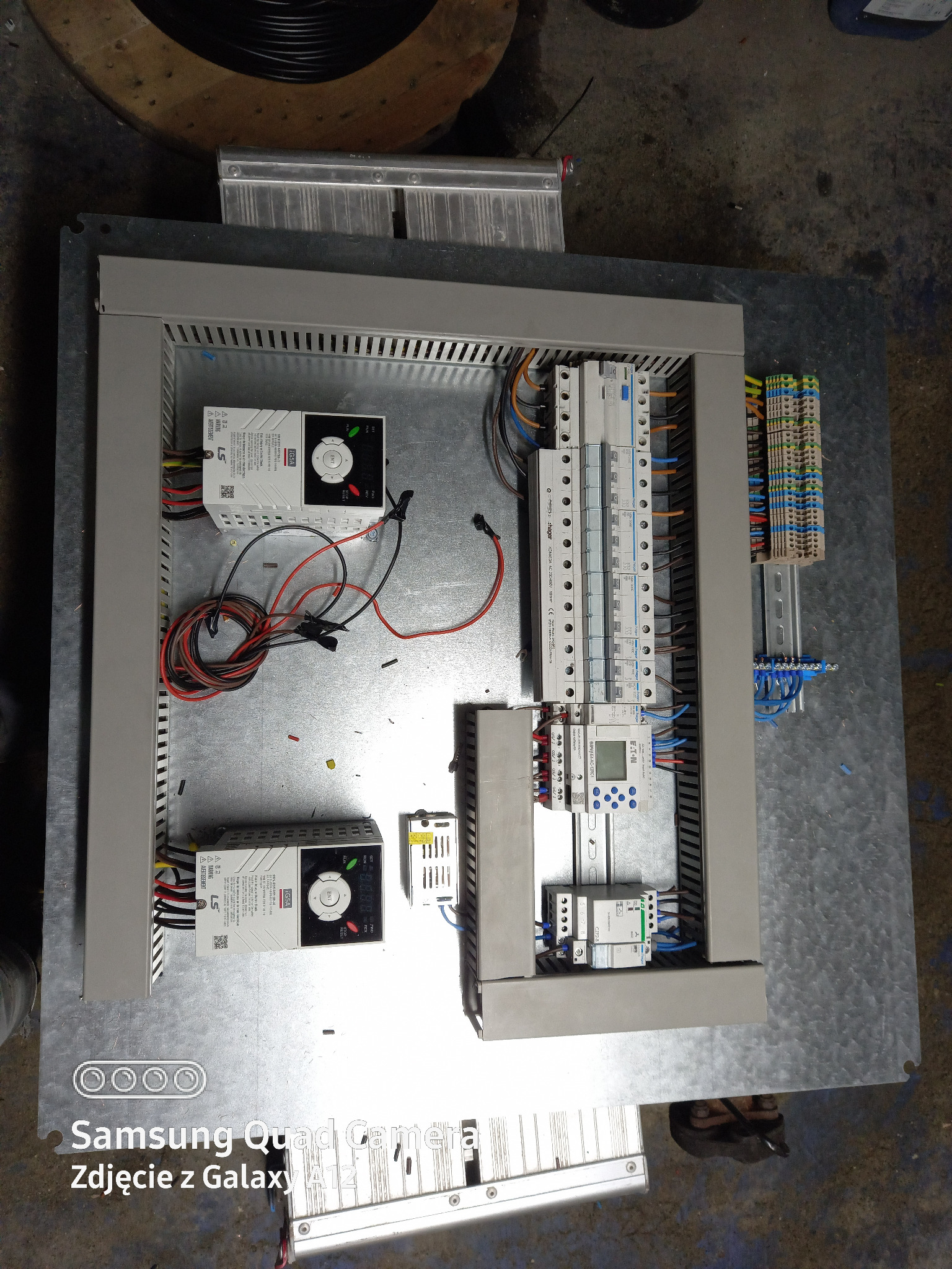 Rozdzielnica elektryczna w trakcie montażu, widoczne zabezpieczenia, sterowniki, przewody i listwy zaciskowe na metalowej płycie montażowej.