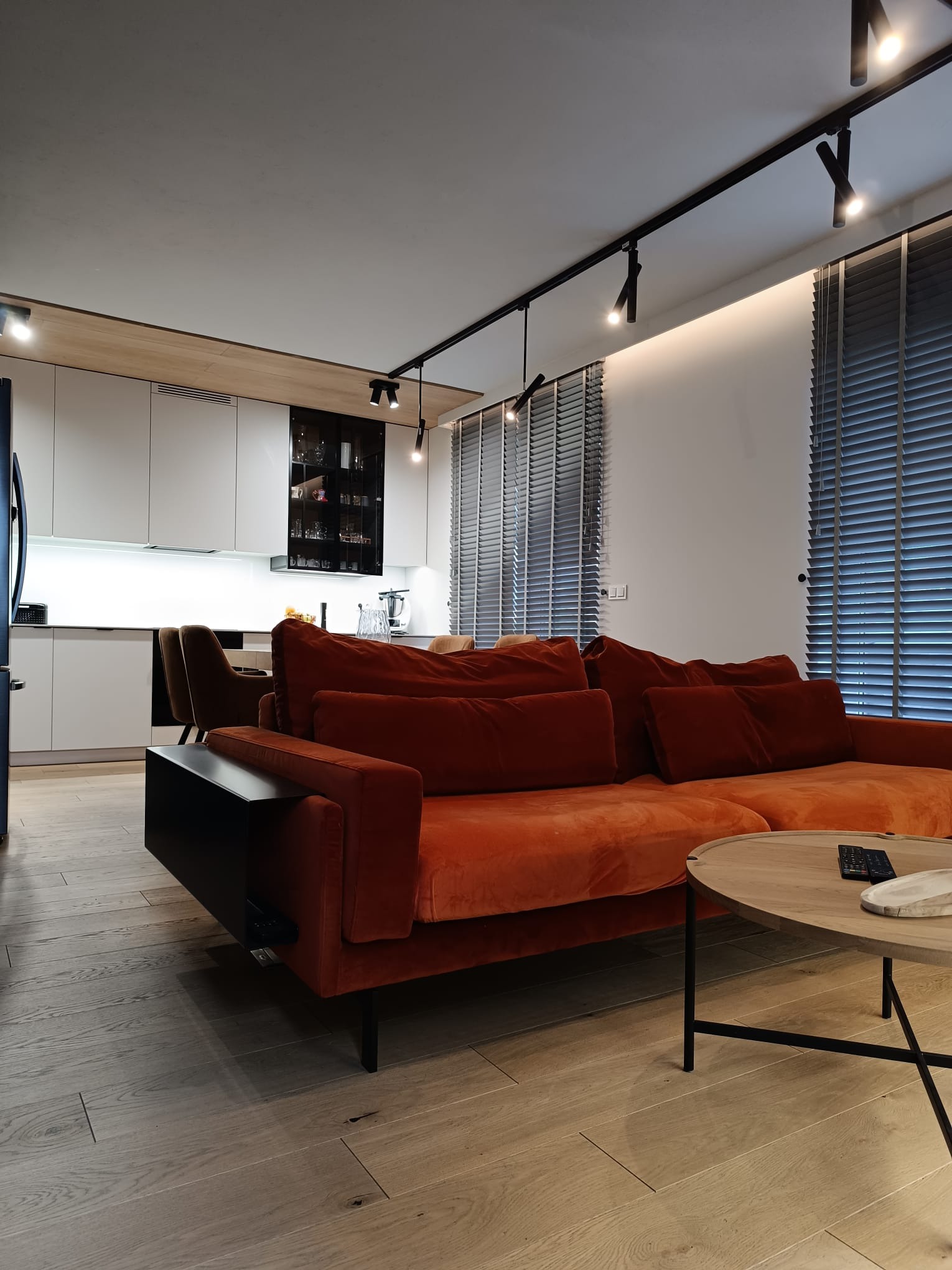 Nowoczesne wnętrze salonu z pomarańczową sofą, okrągłym stolikiem kawowym i minimalistyczną kuchnią w tle, oświetlone reflektorami na szynoprzewodzie.