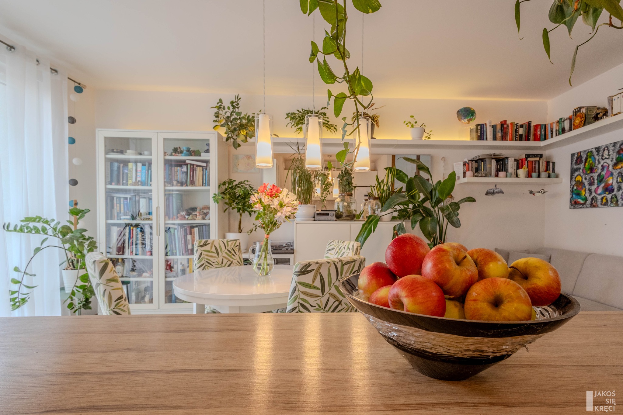 Jasne wnętrze jadalni z okrągłym stołem, dekoracjami roślinnymi i misą jabłek na pierwszym planie, widziane z perspektywy blatu stołu.