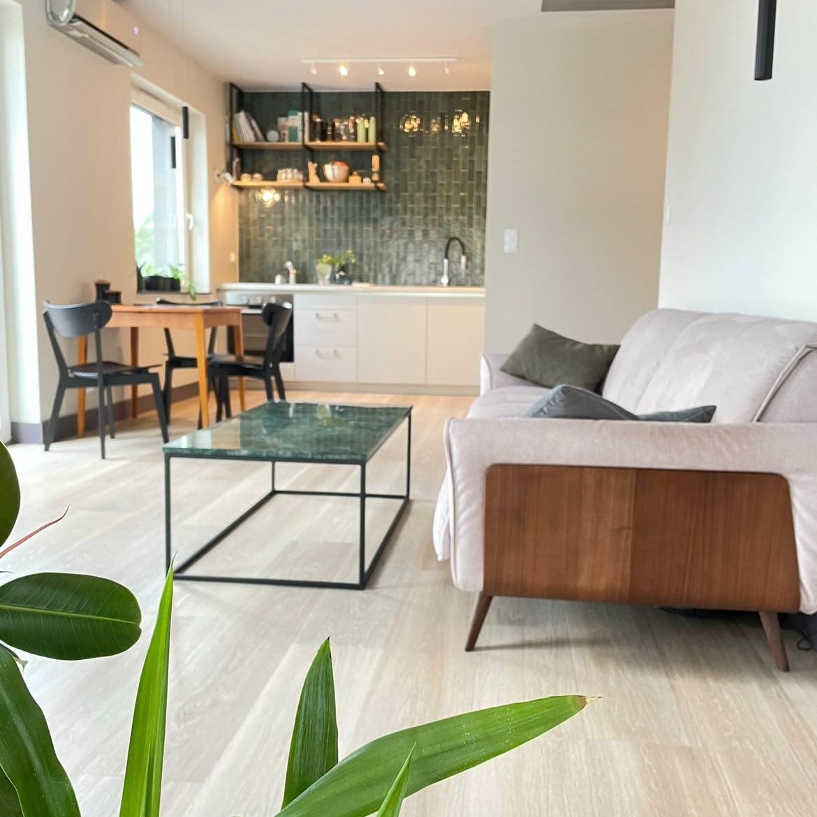 Jasne, nowocześnie urządzone wnętrze mieszkania z otwartą kuchnią, zielonymi płytkami na ścianie i marmurowym stolikiem kawowym na pierwszym planie, z widocznym fragmentem rośliny doniczkowej.