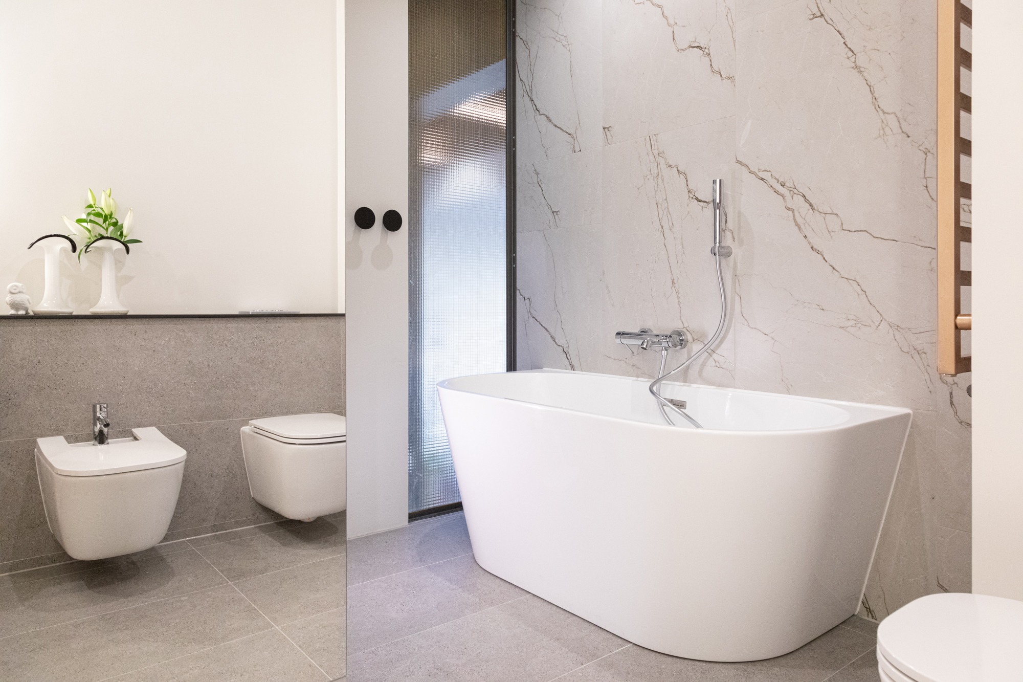 Nowoczesna łazienka z wolnostojącą białą wanną, bidetem i toaletą w minimalistycznym stylu, marmurowe płytki na ścianie, szklana kabina prysznicowa z fakturą, dekoracyjne wazony z kwiatami na półce.