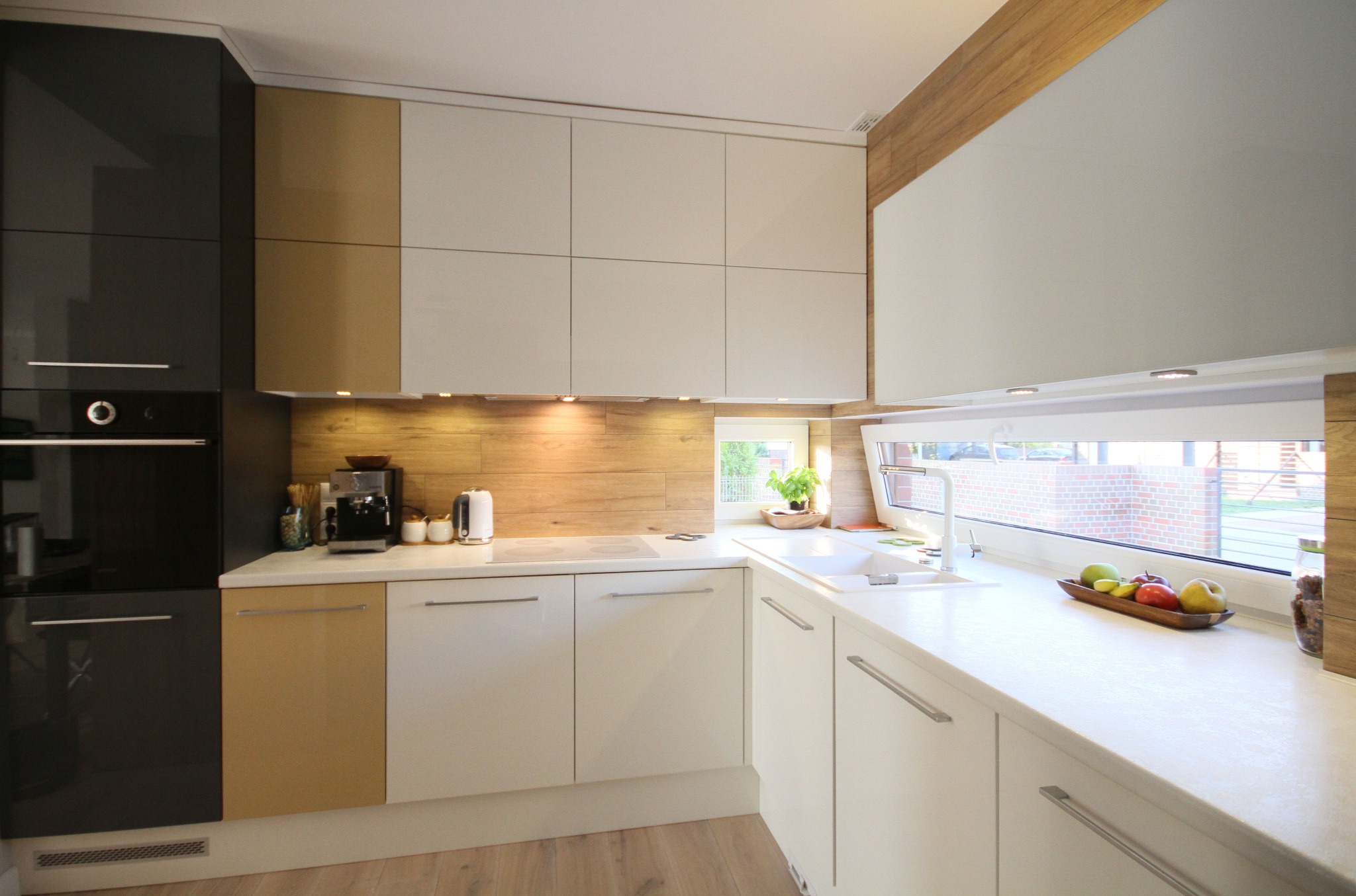 Nowoczesna kuchnia z białymi i brązowymi szafkami, drewnianym panelem nad blatem, widokiem na ogród przez okno i owocami w drewnianej misie.
