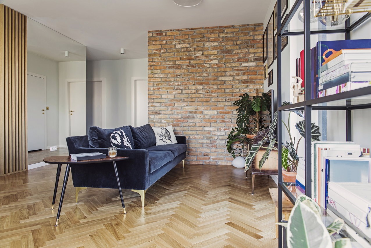 Salon z ceglaną ścianą, parkietem w jodełkę, granatową sofą i roślinami w doniczkach.