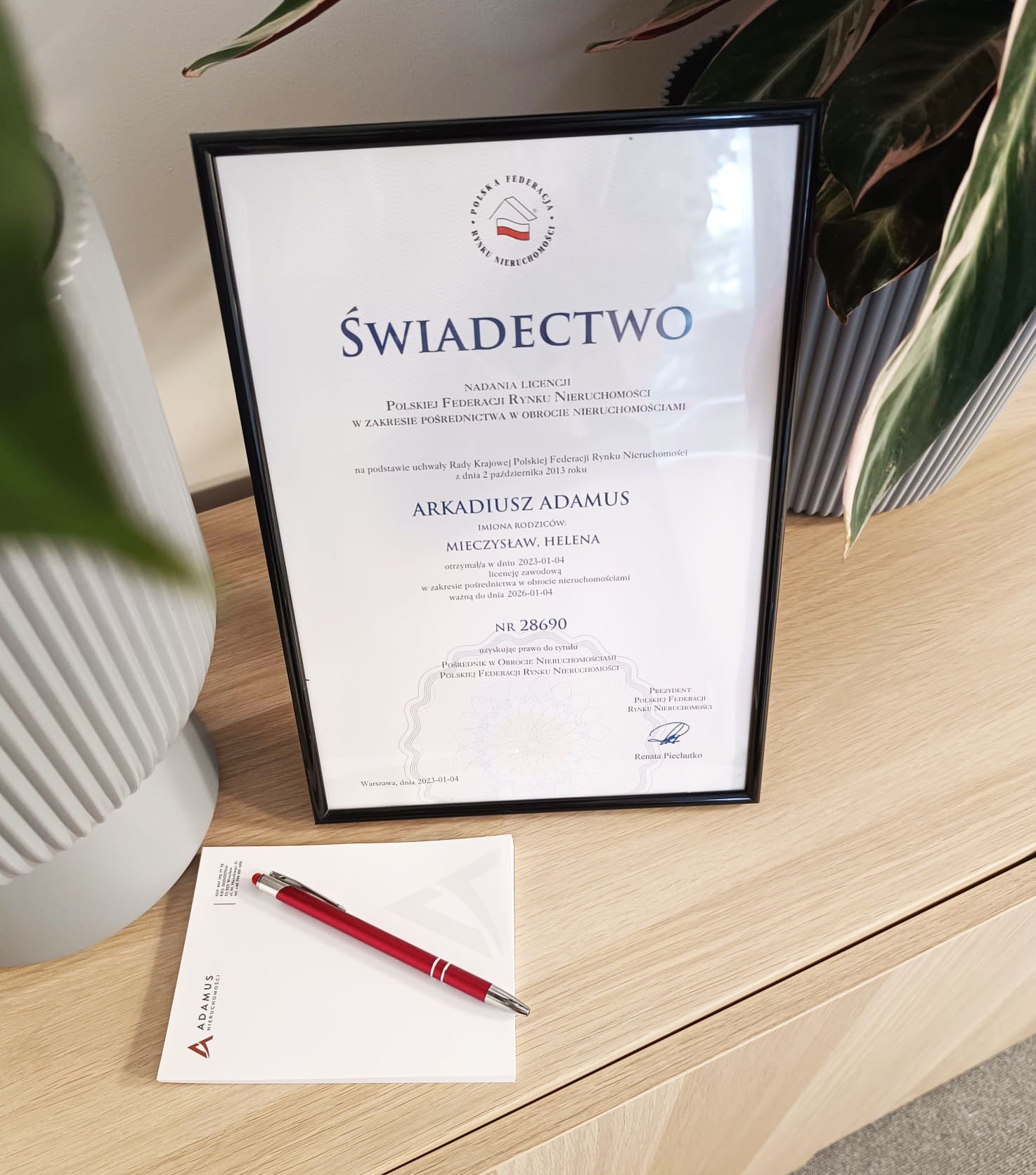 Oprawione świadectwo nadania licencji pośrednika w obrocie nieruchomościami Arkadiusza Adamusa, leżące na biurku obok notatnika firmowego i czerwonego długopisu.
