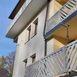 Wielopiętrowy budynek z balkonami po renowacji. Widoczne odnowione balustrady balkonowe o ukośnym wzorze, świeżo pomalowana elewacja oraz rynna spustowa. Na pierwszym planie fragment elewacji...