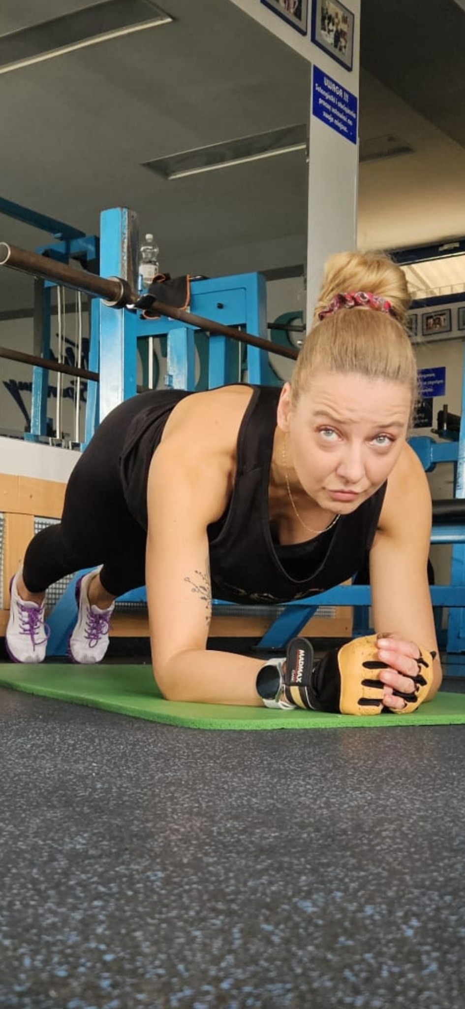 Kobieta w czarnym stroju sportowym wykonuje ćwiczenie plank na zielonej macie w siłowni, w tle widoczny sprzęt do ćwiczeń siłowych i tablica informacyjna.