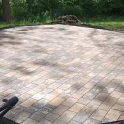 Nowo ułożony, wielobarwny chodnik z kostki brukowej w kształcie wielokąta, otoczony ziemią i trawnikiem, z widocznym fragmentem narzędzia brukarskiego w rogu.