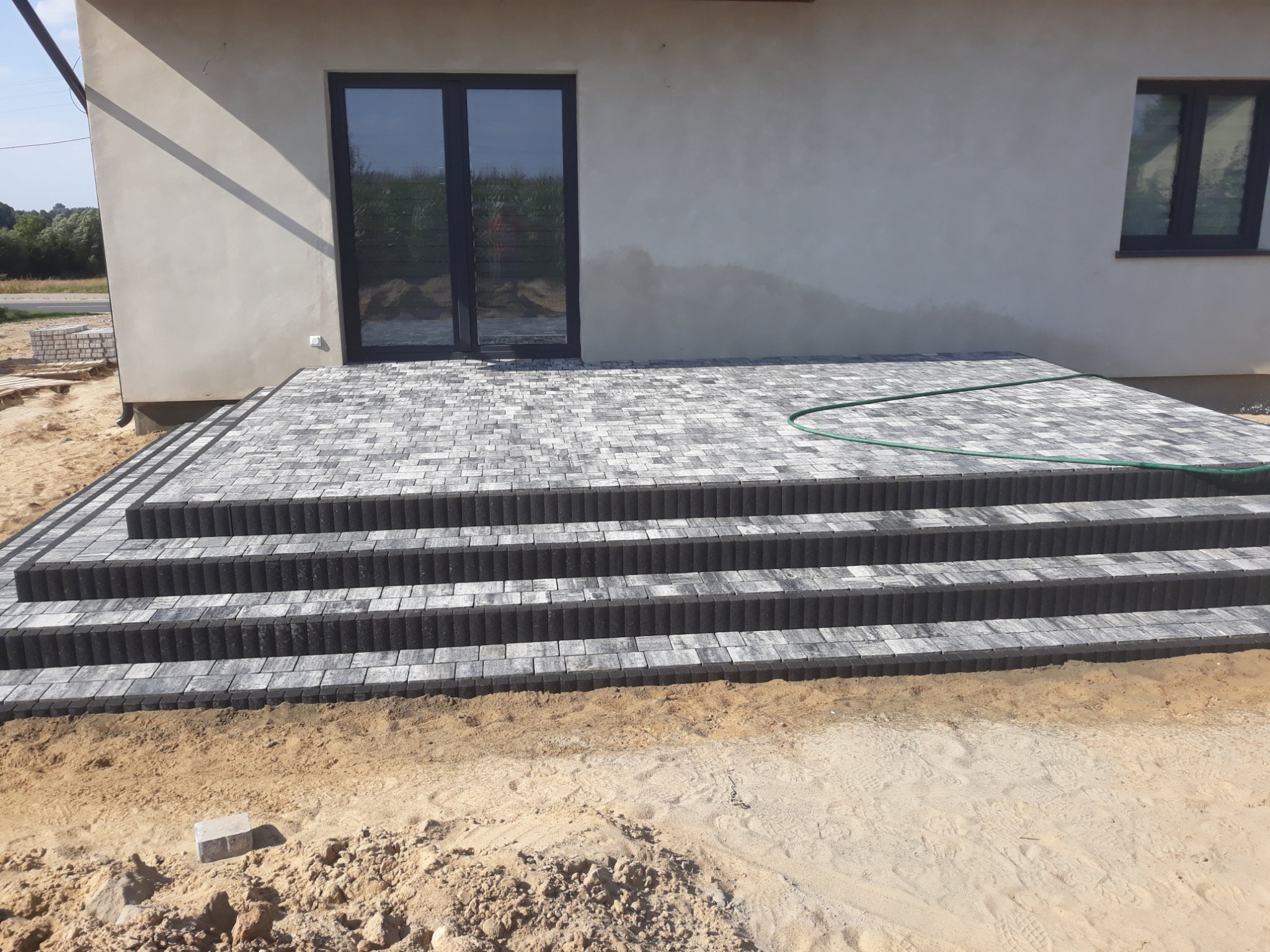 Wykończony taras z kostki brukowej w odcieniach szarości, prowadzący do drzwi balkonowych nowoczesnego domu. Widoczna zielona rura na tarasie. W tle niedokończone elementy krajobrazu.