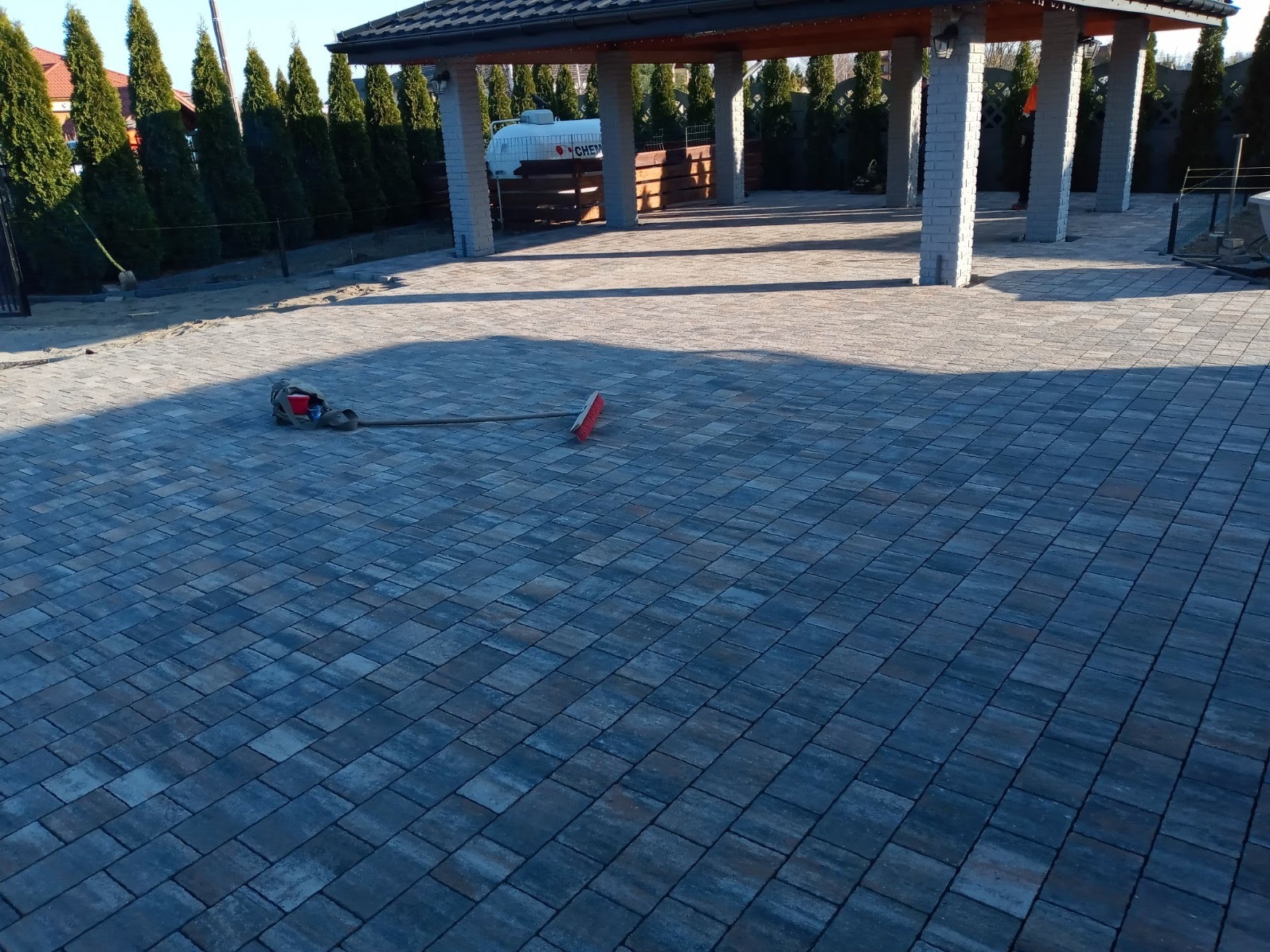 Nowo ułożona kostka brukowa w odcieniach szarości i brązu na posesji z zadaszeniem i tujami w tle, obok leży miotła i torba narzędziowa.