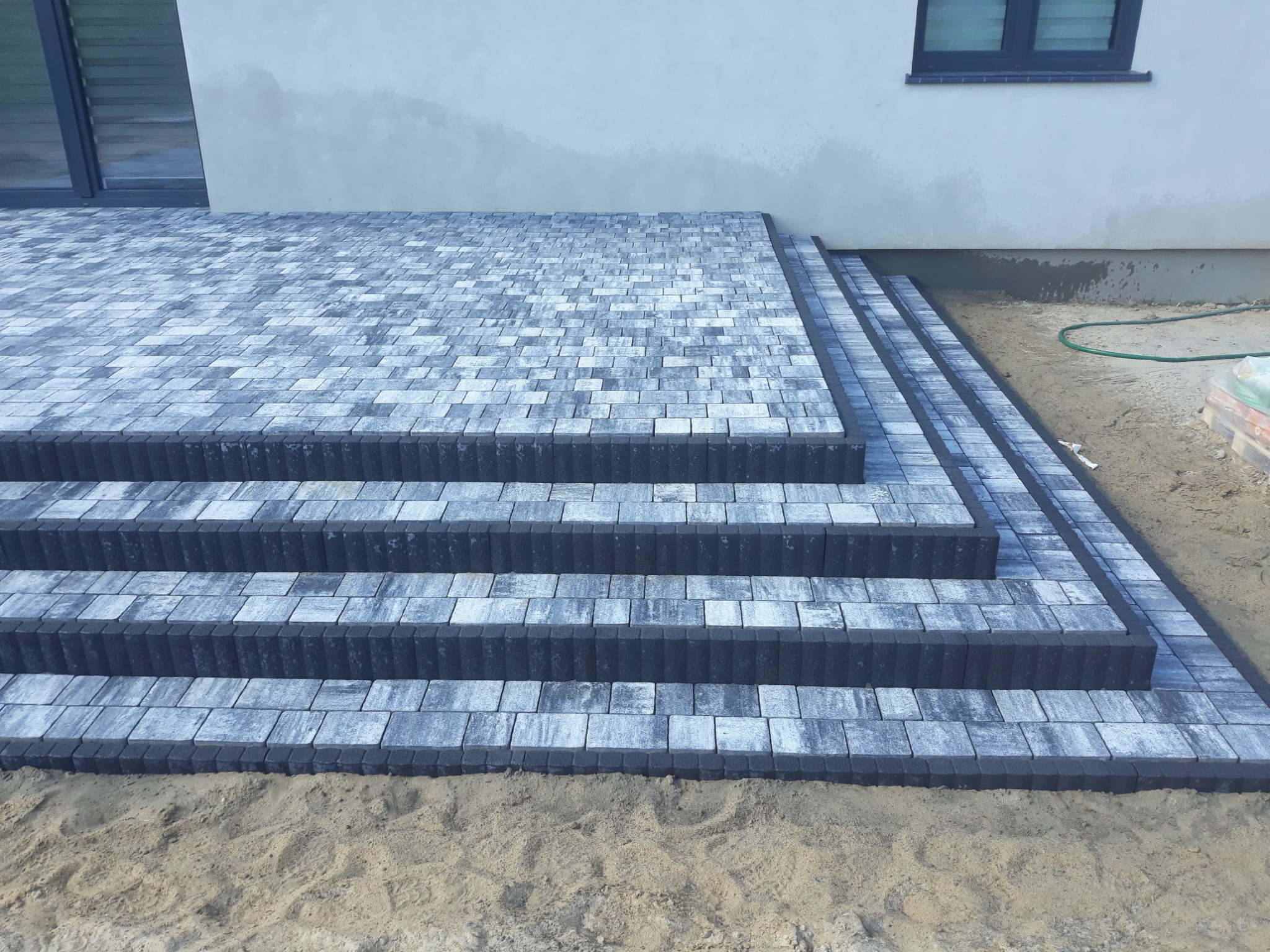 Schody zewnętrzne wyłożone kostką brukową w odcieniach szarości, prowadzące do budynku z jasną elewacją. Widoczny fragment trawnika i piasek.