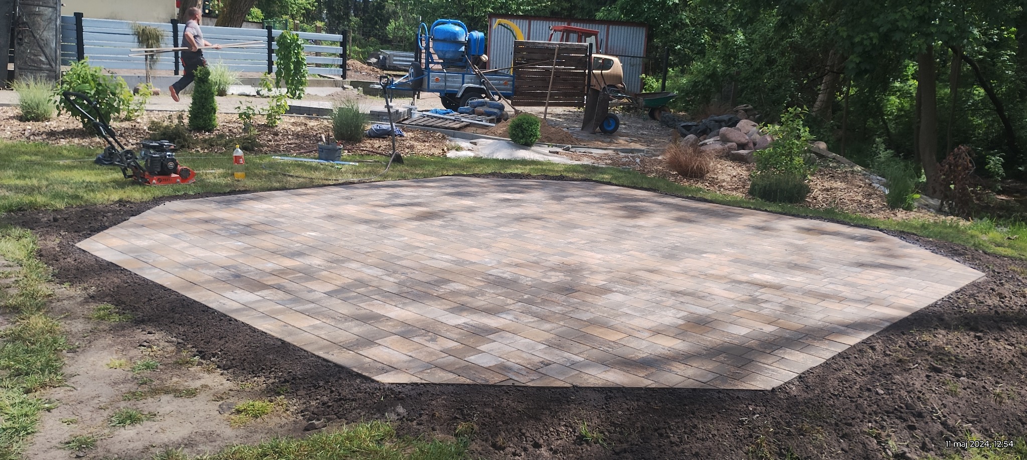 Ukończona, wielobarwna nawierzchnia z kostki brukowej o nieregularnym kształcie na trawniku, z widocznymi narzędziami i materiałami budowlanymi w tle.
