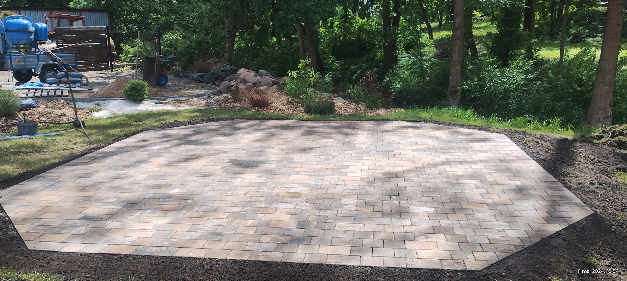 Wykończony, wielobarwny plac z kostki brukowej ułożonej w regularny wzór, otoczony świeżo obsypaną ziemią i zielenią ogrodu; w tle widoczny sprzęt budowlany i drzewa.