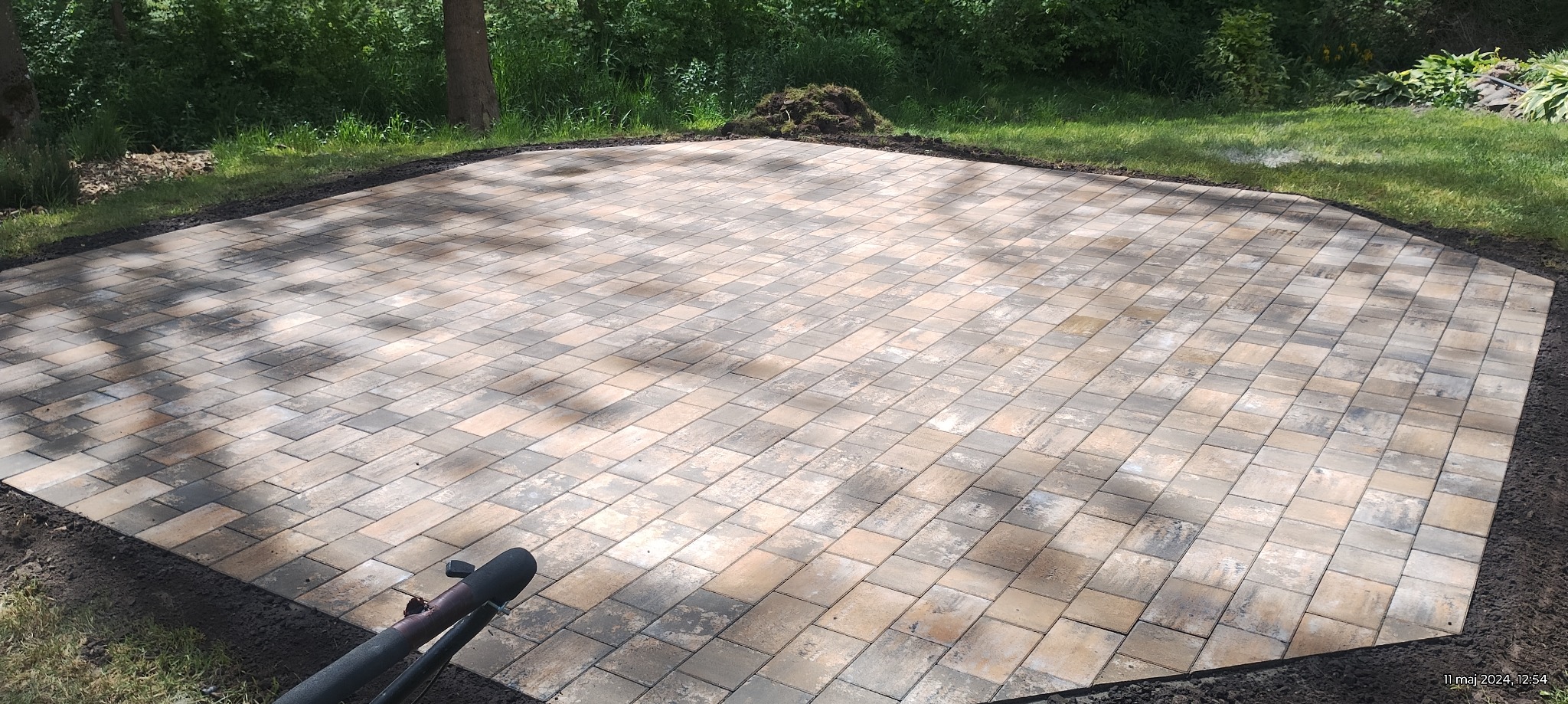 Nowo ułożony, wielobarwny chodnik z kostki brukowej w kształcie wielokąta, otoczony ziemią i trawnikiem, z widocznym fragmentem narzędzia brukarskiego w rogu.