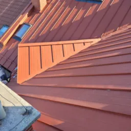 Czerwony, metalowy dach z oknami dachowymi i kominem, widok z góry, fragment blachy z pionowymi panelami.