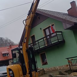 Usługi Koparką Łukasz Bereśniewicz - Żółta koparka CAT 303.5E z podniesioną łyżką na brukowanej nawierzchni, w tle zielony dom z balkonem i polską flagą.