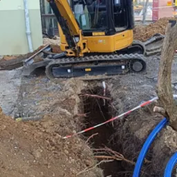 Żółta mini koparka CAT obok wykopu z widocznymi korzeniami, zabezpieczonego taśmą ostrzegawczą i z niebieskimi rurami w środku.
