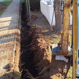 Usługi Koparką Łukasz Bereśniewicz - Długi, wąski wykop w ziemi, widoczny z perspektywy operatora koparki, obok chodnika i bloku mieszkalnego z adresem na ścianie, odsłonięte korzenie drzew w wykopie.