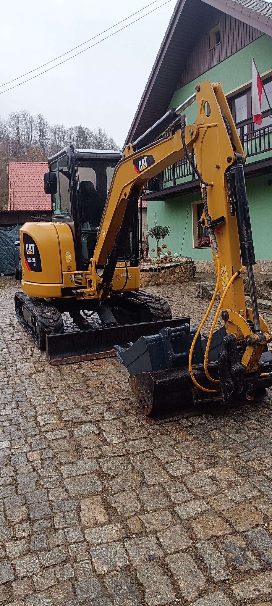 Żółta minikoparka CAT 303.5E na brukowanej nawierzchni, z widocznym łyżką do kopania, budynek mieszkalny w tle z powiewającą polską flagą.