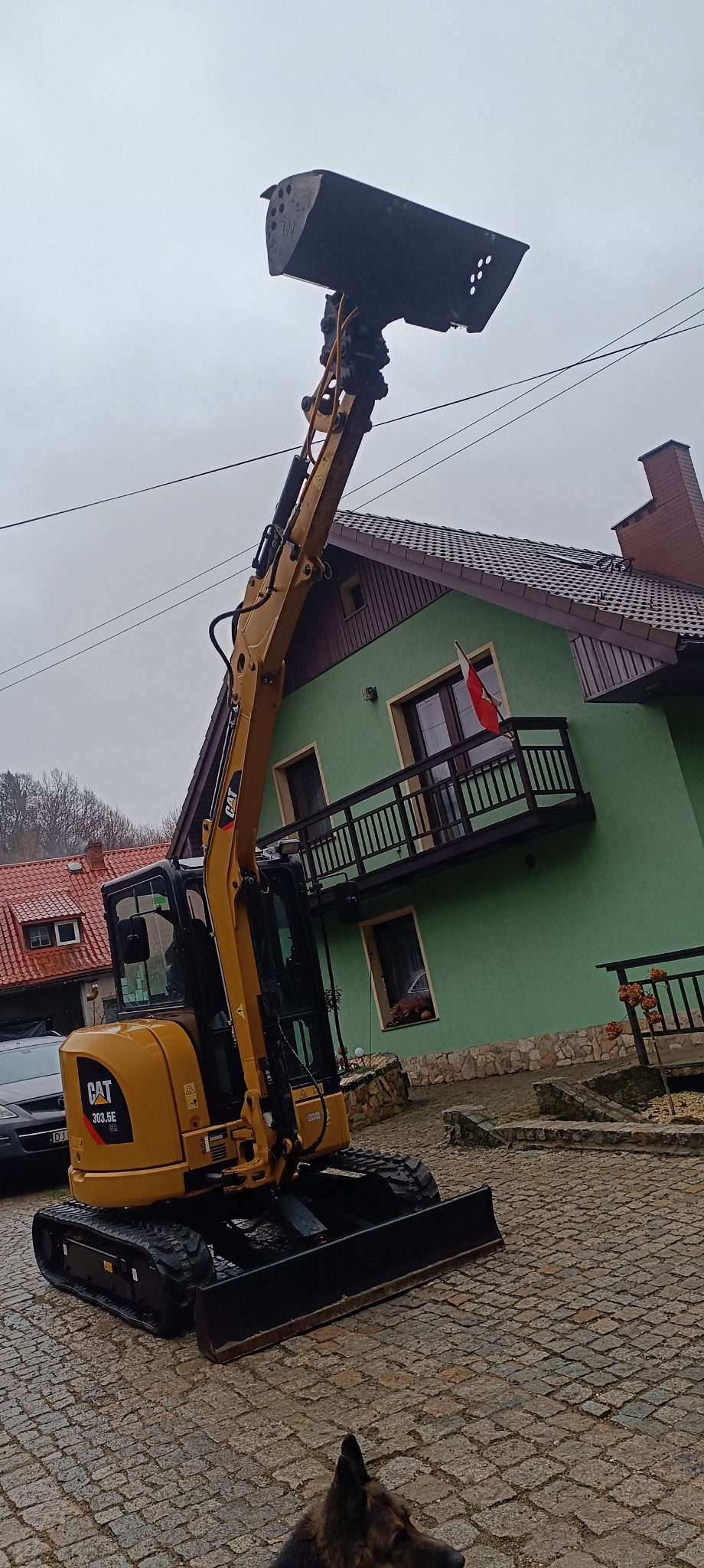 Żółta koparka CAT 303.5E z podniesioną łyżką na brukowanej nawierzchni, w tle zielony dom z balkonem i polską flagą.