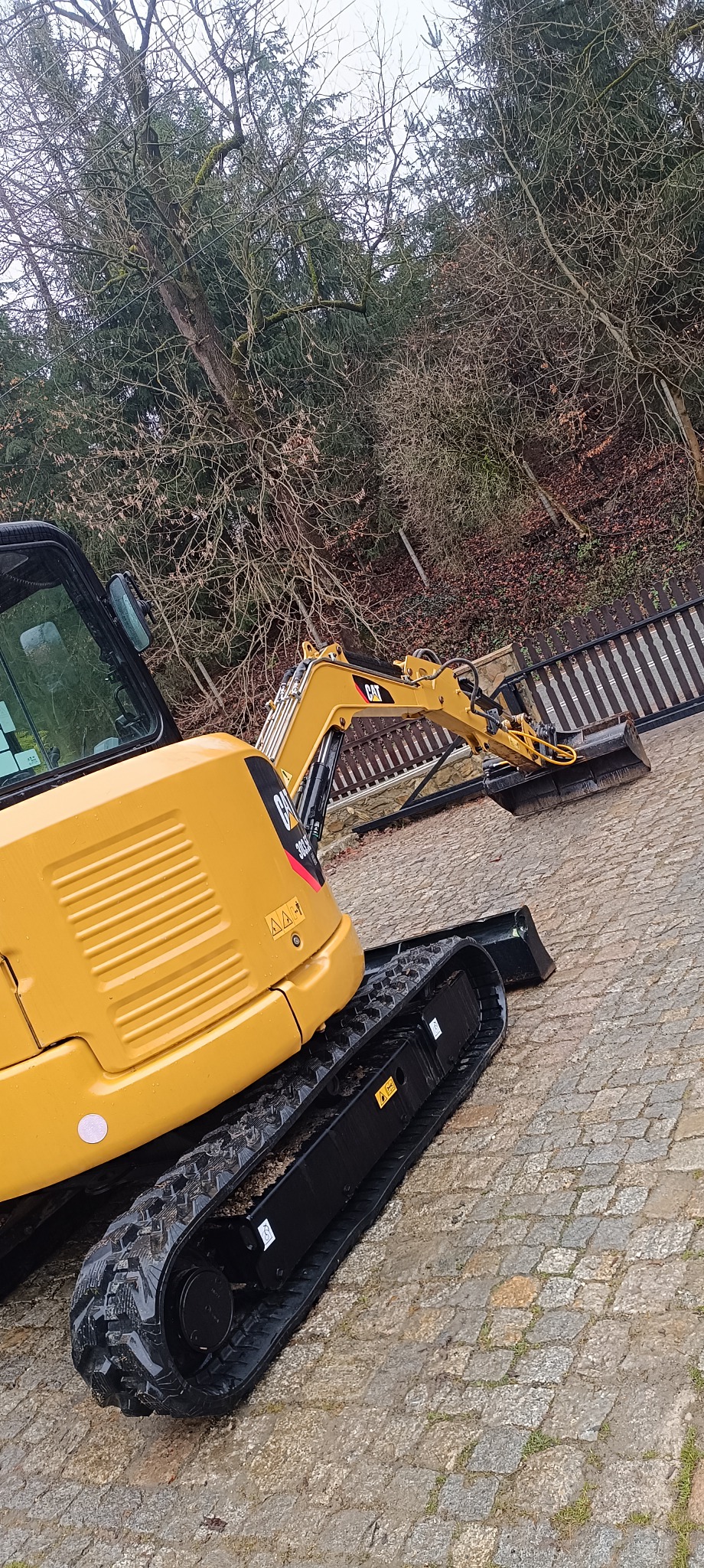 Żółta minikoparka CAT 303CR na brukowanym podłożu, z widocznymi gąsienicami i łyżką skierowaną w stronę drewnianego ogrodzenia, drzewa w tle.