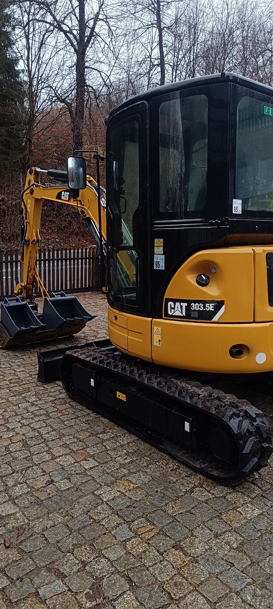 Żółto-czarna mini koparka CAT 303.5E CR na kostce brukowej, widoczne łyżki koparki, kabina z naklejkami informacyjnymi, w tle drewniany płot i drzewa.