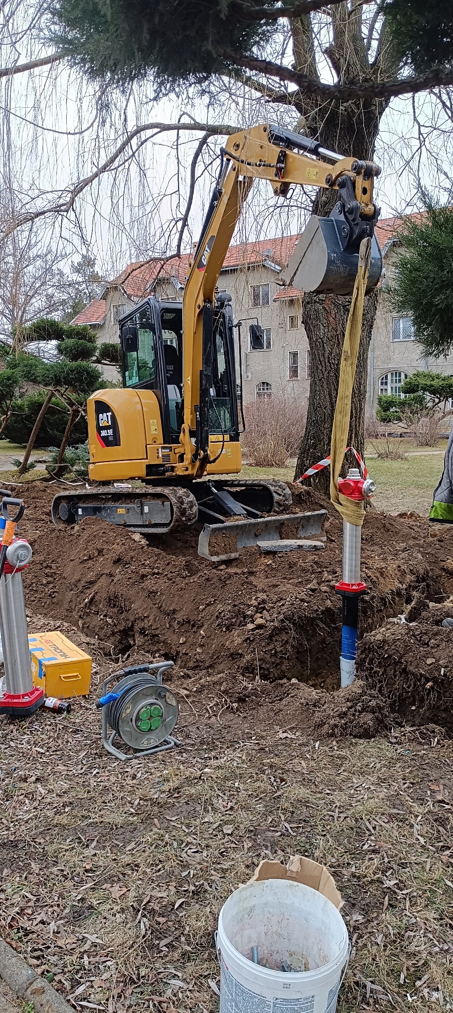 Żółta minikoparka CAT 303.5E w trakcie wykopu pod instalację hydrantu przeciwpożarowego zabezpieczonego pasem transportowym, widoczne narzędzia i skrzynka elektryczna, budynek w tle.