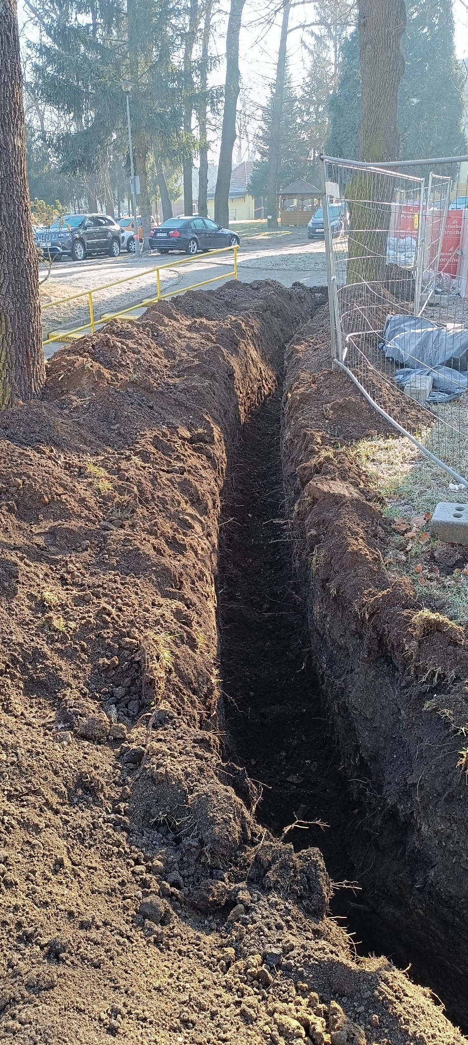 Długi, wąski rów wykopany w ziemi, zabezpieczony siatką ogrodzeniową i żółtą barierką, widoczne drzewa w tle.