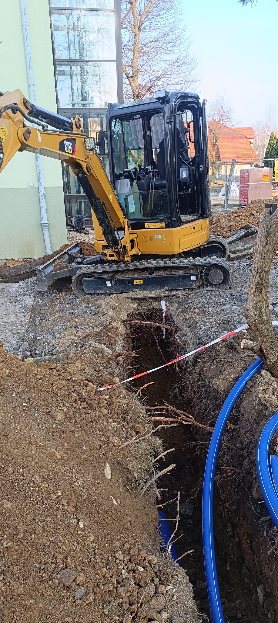 Żółta mini koparka CAT obok wykopu z widocznymi korzeniami, zabezpieczonego taśmą ostrzegawczą i z niebieskimi rurami w środku.