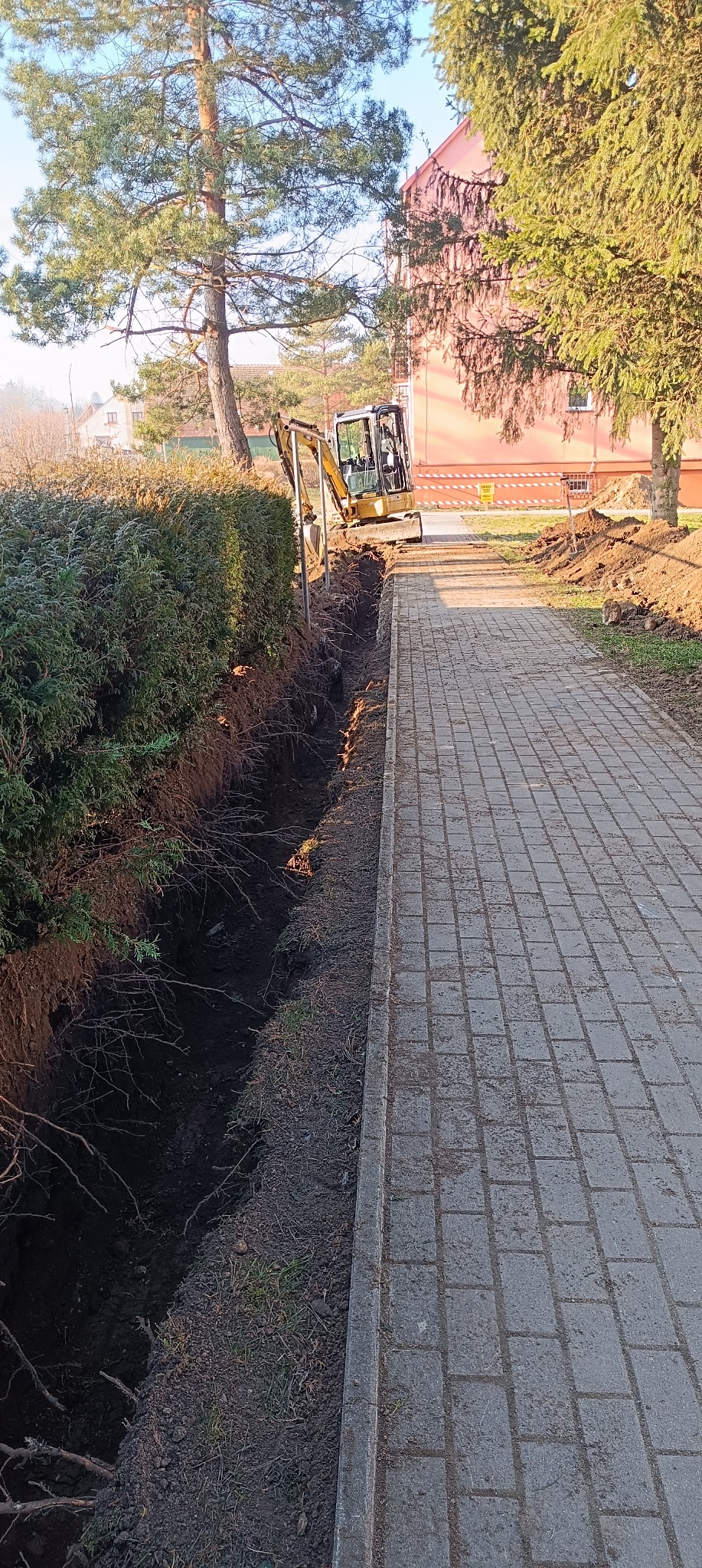 Wąski wykop wzdłuż chodnika z kostki brukowej, wykonany przy użyciu małej koparki, obok żywopłotu i drzew, z budynkiem w tle.