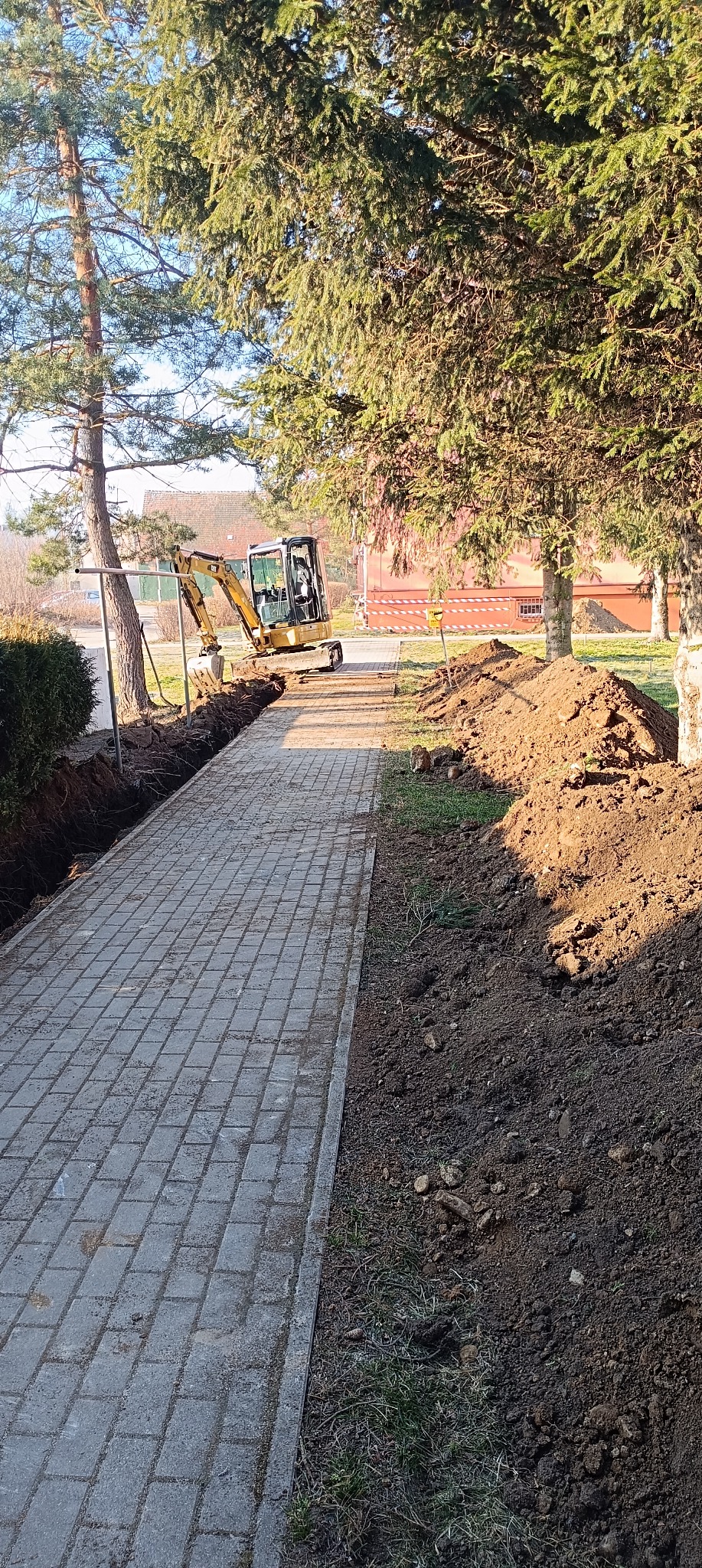 Żółta minikoparka wykonuje wąski wykop wzdłuż ścieżki z kostki brukowej, pryzmy ziemi po obu stronach wykopu, drzewa w tle.