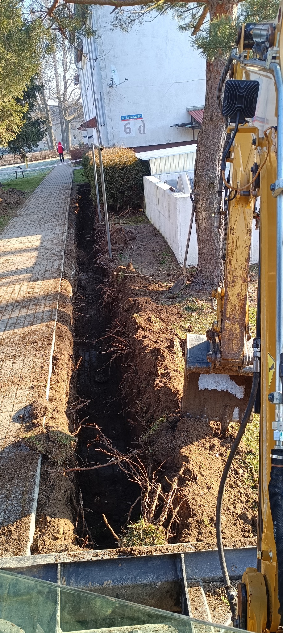 Długi, wąski wykop w ziemi, widoczny z perspektywy operatora koparki, obok chodnika i bloku mieszkalnego z adresem na ścianie, odsłonięte korzenie drzew w wykopie.