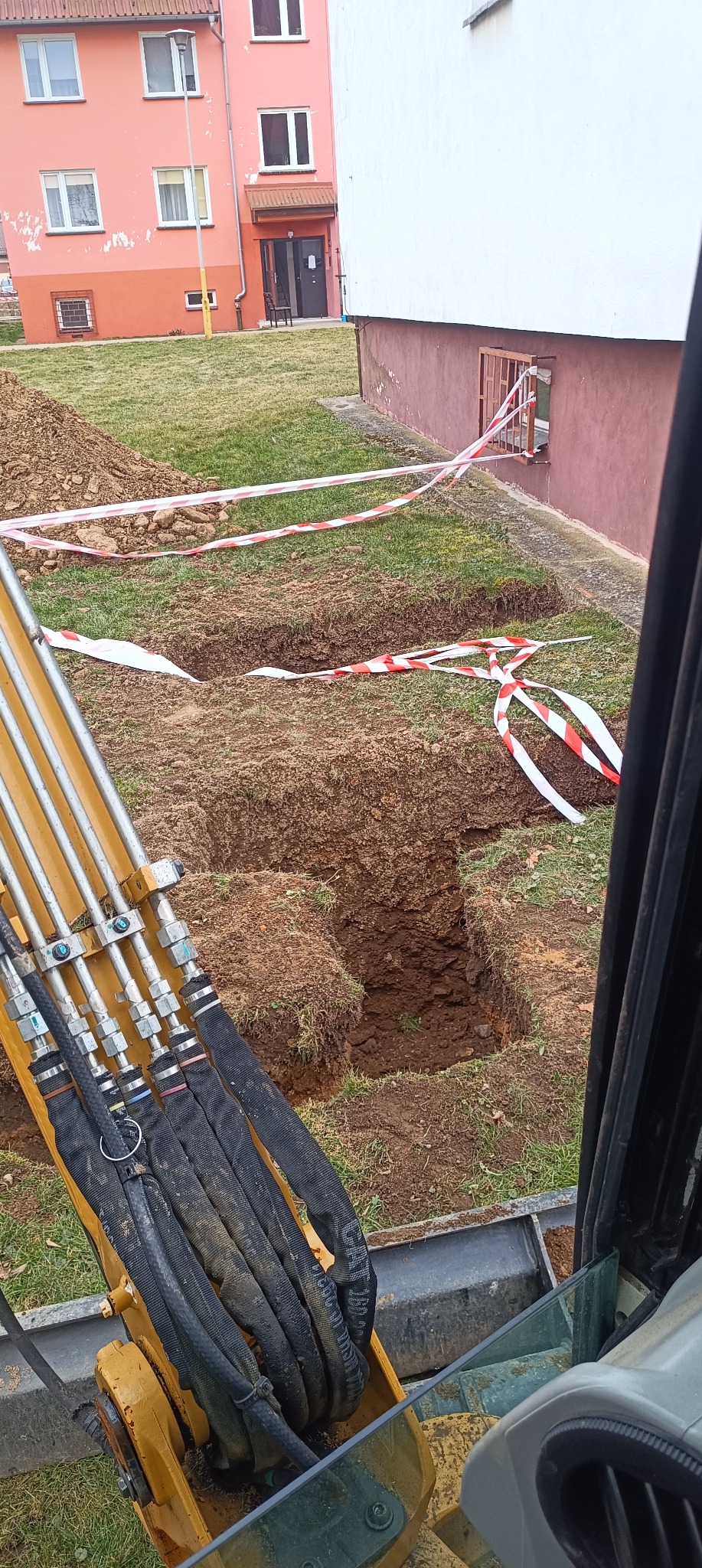Wykop pod instalacje wykonany koparką, zabezpieczony taśmą ostrzegawczą na trawniku przed budynkiem mieszkalnym.