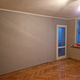 Adam Piotrowski - Pomieszczenie po odświeżeniu ścian szarą farbą, widoczny parkiet ułożony w jodełkę, okno z widokiem na drzewa i akcesoria malarskie na parapecie.