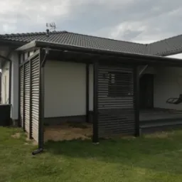 Nowoczesna konstrukcja z drewnianych paneli osłaniająca taras przy domu z ciemną dachówką, widoczny fragment ogrodu i wiszące krzesło.