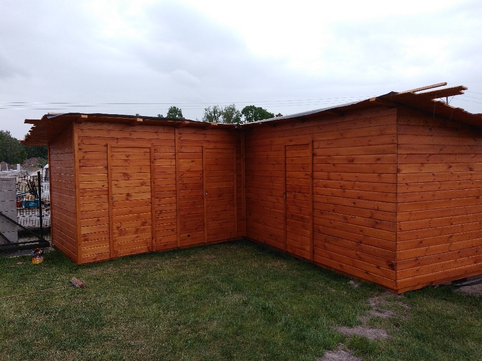 Brązowa, drewniana konstrukcja z dwiema parami drzwi na trawniku, widoczna w pochmurny dzień, z elementami w trakcie montażu dachu.