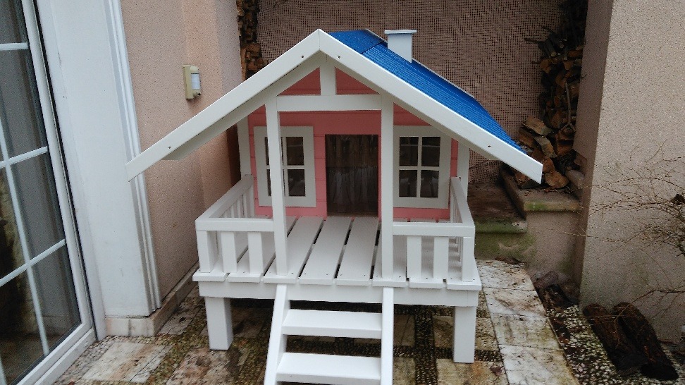 Mały, pomalowany na różowo i biało domek z niebieskim dachem, imitujący budynek mieszkalny, z miniaturowym tarasem i schodkami, ustawiony na zewnątrz na wyłożonym kamieniami podłożu.