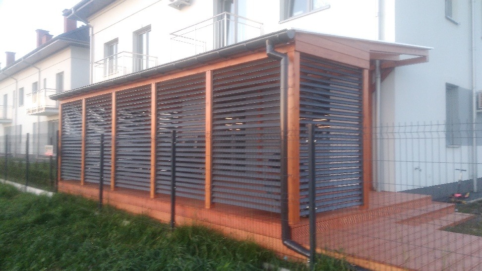 Zadaszona konstrukcja z drewnianych słupów i szarych, poziomych żaluzji, przylegająca do budynku, z widocznymi schodami wejściowymi i rynną odprowadzającą wodę.