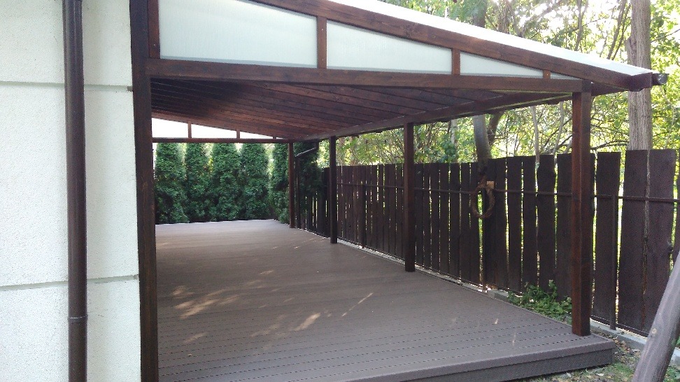 Zadaszony taras z drewnianą konstrukcją, brązową podłogą z desek kompozytowych i ciemnym drewnianym ogrodzeniem, widok na drzewa w tle.