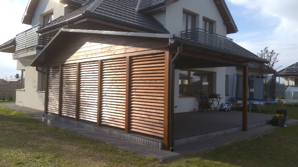 Zadaszona przestrzeń przy domu z drewnianą konstrukcją i poziomymi żaluzjami, z widocznym tarasem, meblami ogrodowymi i trampoliną w tle.