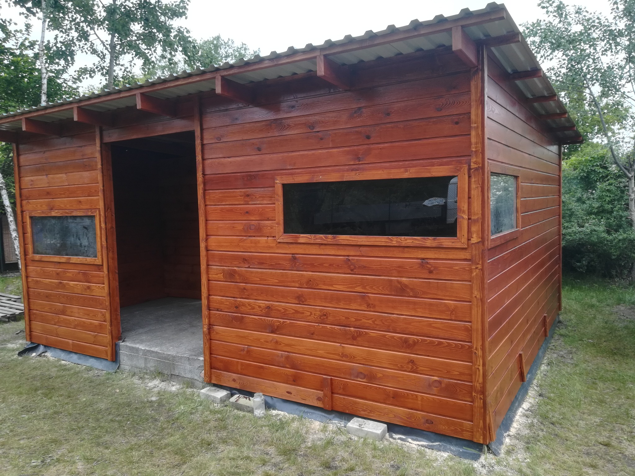 Brązowa, drewniana konstrukcja z metalowym dachem, z otwartym wejściem i oknami, postawiona na betonowych bloczkach na trawniku.