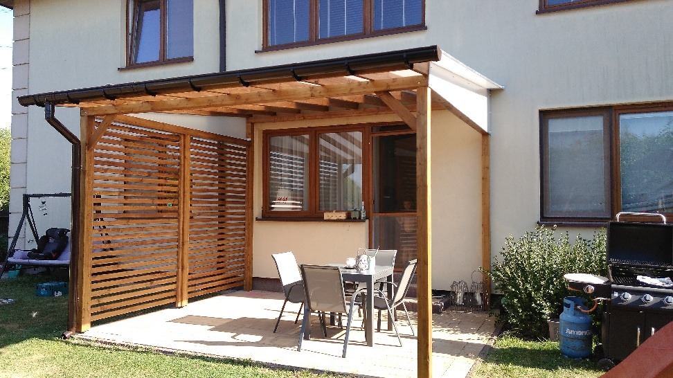 Zadaszona konstrukcja z drewnianych belek, tworząca zacienione miejsce do siedzenia z ażurową ścianką, stolik z krzesłami i grill na zewnątrz budynku.