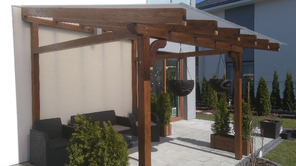 Zadaszona drewniana konstrukcja przylegająca do ściany budynku, z meblami ogrodowymi i wiszącymi donicami, otoczona zielenią.