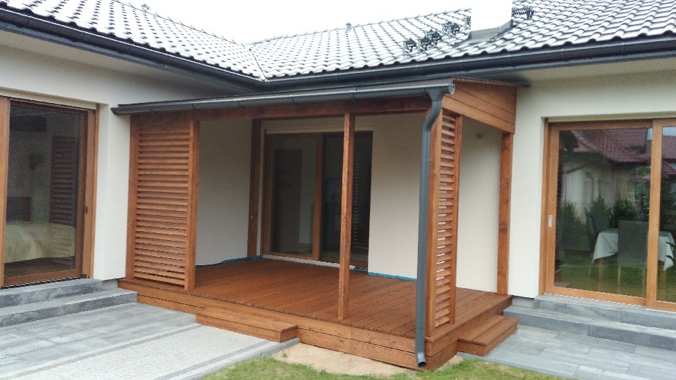 Zadaszony taras przylegający do domu z drewnianą podłogą i ażurowymi ściankami bocznymi, widoczne okna przesuwne i fragment dachu.