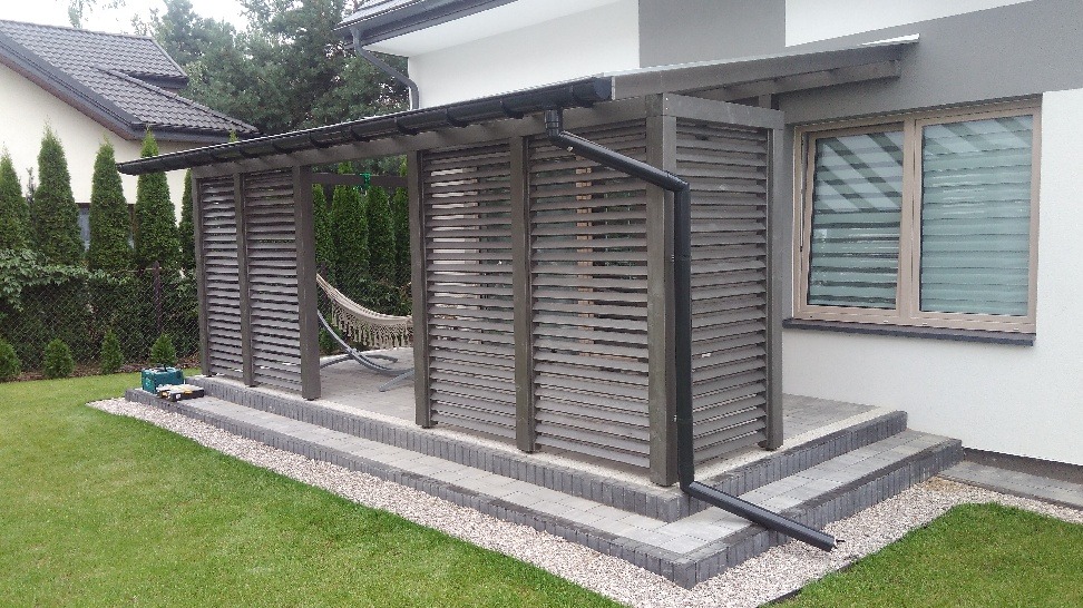 Zadaszona konstrukcja z drewnianymi żaluzjami, prawdopodobnie na tarasie, z widocznym hamakiem wewnątrz, usytuowana przy budynku z oknem i zielonym trawnikiem.