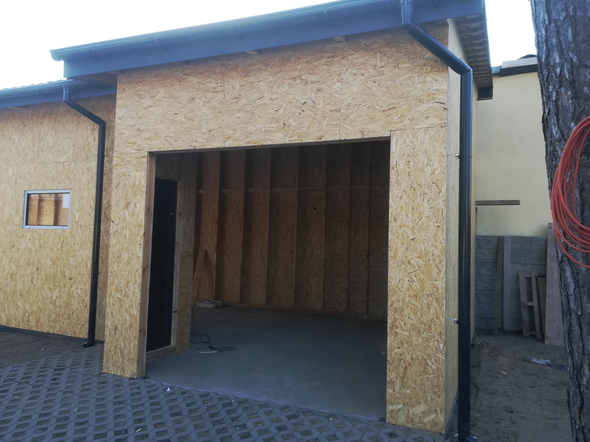 Surowa konstrukcja garażu z płyty OSB, widoczne rynny i betonowa posadzka, częściowo wykończony budynek na zewnątrz z ułożoną kostką ażurową.